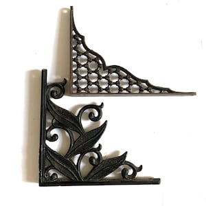 May include: Two L-shaped metal shelf brackets. The top bracket is dark brown with a repeating circular lattice pattern. The bottom bracket is black with a decorative leaf and scrollwork design. Both brackets feature mounting holes along the edges.