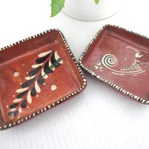 May include: Two square, terracotta-colored ceramic dishes. One dish features a black, white, and green leaf design. The other has a snail and swirl design. Both have white dotted edges.
