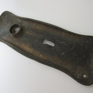 May include: A vintage brass door plate with a keyhole and a round hole for a knob. The plate has a slightly curved shape and a worn, aged finish.
