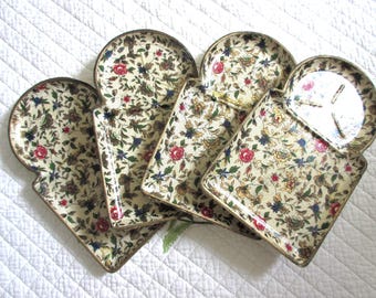 May include: A set of four metal trays with a cream-colored background featuring a repeating floral pattern of red roses, blue flowers, and green leaves. Each tray has a rounded top section and a rectangular base with a metallic gold-toned rim.