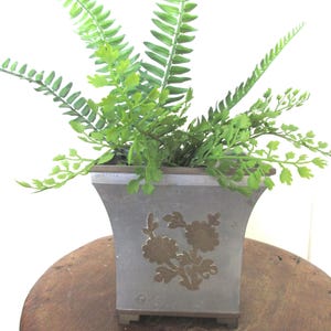 Può includere: Un vaso quadrato in metallo grigio con un intarsio floreale color ottone sul pannello frontale. Il vaso presenta un bordo superiore svasato e una base piccola, e contiene una felce artificiale verde con molteplici fronde.