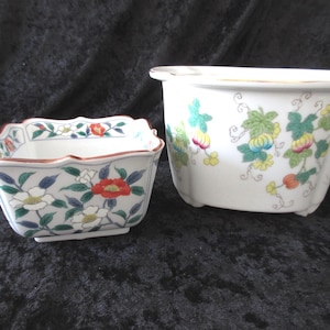 May include: A square ceramic bowl with a scalloped rim featuring a floral pattern in red, white, and green, alongside a larger white ceramic planter decorated with a colorful vine and pumpkin motif, both featuring gold-toned trim on the edges.