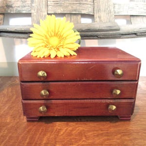May include: A small, rectangular wooden chest with a reddish-brown finish featuring three horizontal drawers. Each drawer is equipped with a single, small, conical brass-colored knob. A bright yellow artificial flower rests on the top surface.