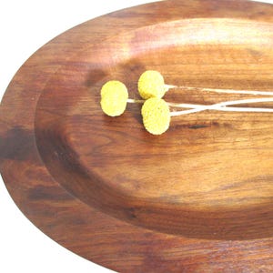 May include: A wooden serving tray with a dark brown finish. Three yellow, spherical decorative flowers with white stems are arranged on the tray. The tray has a smooth, polished surface and a shallow, oval shape.