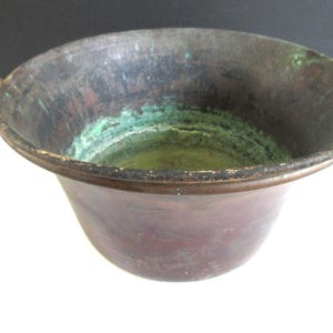 Op de afbeelding: Een antieke koperen ketel met een afgeronde body en een brede, licht uitlopende rand. De ketel heeft twee kleine handvatten en vertoont tekenen van ouderdom met een patina van groene en bruine tinten. De binnenkant heeft een groenachtige tint.