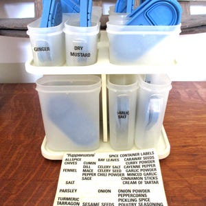 Könnte beinhalten: Ein Vintage-Gewürzregal von Tupperware mit transparenten Behältern und blauen Deckeln. Das Regal enthält beschriftete Behälter für Gewürze wie Ingwer, Trocken-Senf und Knoblauchsalz. Ein Etikett listet verschiedene Gewürznamen auf.