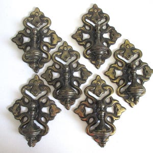 May include: A set of six brass drawer pulls, each featuring an ornate, symmetrical backplate with intricate scrollwork and a central drop handle. The metal has a darkened, aged patina with visible highlights on the raised decorative edges.