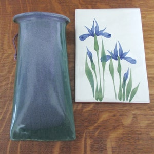 Può includere: Un vaso in ceramica blu e verde con un manico in corda marrone, accanto a una piastrella in ceramica bianca con un disegno dipinto a mano di iris blu.