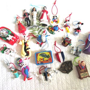 May include: A collection of various hanging Christmas ornaments featuring cartoon characters, figures, and a small rectangular tin with a graphic illustration. The assortment includes Disney characters, a Santa figure, and other colorful holiday decorations.