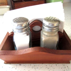 May include: A wooden salt and pepper shaker set with a built-in napkin holder. The set includes two glass shakers with silver lids, one filled with salt and the other with pepper. The wooden holder is stained a warm brown.