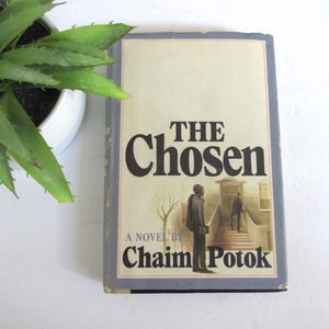 May include: A hardcover book titled "THE Chosen" by Chaim Potok. The book cover features a muted color palette with a drawing of two figures on a porch. A green plant in a white pot is in the upper left corner.