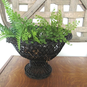 May include: A black, woven wire basket with a pedestal base, filled with green fern plants. The basket has a decorative, open weave design. The basket is sitting on a wooden surface.