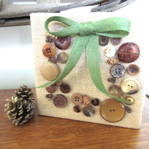 May include: A square canvas covered in burlap, adorned with various brown buttons and a green ribbon bow. A pinecone sits to the left. The buttons are arranged in a decorative pattern.