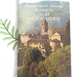 May include: A book titled "Sonnets From The Portuguese" by Elizabeth Barrett Browning. The cover features a scenic landscape with a tower and buildings, and lush green trees. The book is resting on a white surface with a green embroidered leaf.