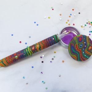 Könnte beinhalten: Eine farbenfrohe Rauchpfeife mit passendem rundem Deckel und einem kleinen Behälter mit violetter Flüssigkeit. Die Pfeife und der Deckel weisen eine lebendige Mischung aus Farben auf, darunter Rot, Orange, Grün und Blau, mit funkelnden Akzenten.