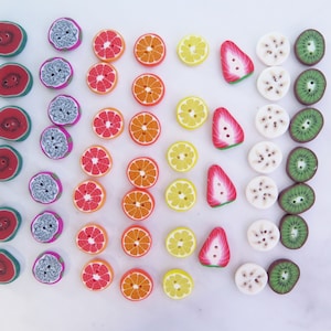 Op de afbeelding: Assortiment van kleurrijke fruitvormige knopen, waaronder watermeloen, kiwi, citroen, sinaasappel, aardbei en ananas. De knopen zijn gerangschikt in rijen per kleur.