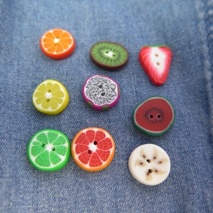 Könnte beinhalten: Sortiment an bunten, fruchtförmigen Knöpfen, darunter eine Wassermelone, eine Erdbeere, eine Kiwi, eine Zitrone, eine Limette, eine Grapefruit und eine Drachenfrucht. Die Knöpfe sind auf einem blauen Jeansstoff angeordnet.