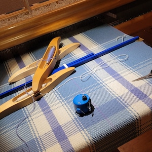 以下が含まれることがあります： 青と白のチェック柄の生地が張られた木製の織機。青い糸と白い糸が入った2つの木製シャトルが生地の上にあります。青いボビンワインダーと青い棒も存在します。