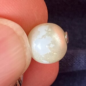 May include: A round, off-white pearl bead featuring a distinct, irregular white map-like pattern on its surface. The bead is spherical with a smooth, lustrous finish and a small metallic component visible on the right side of the pearl.