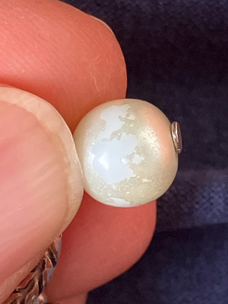 May include: A round, off-white pearl bead featuring a distinct, irregular white map-like pattern on its surface. The bead is spherical with a smooth, lustrous finish and a small metallic component visible on the right side of the pearl.