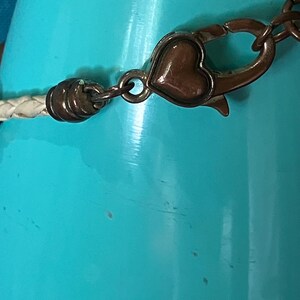 May include: A white braided cord bracelet featuring a metallic bronze-toned lobster claw clasp. The clasp mechanism is accented with a small, smooth, heart-shaped detail in a matching bronze finish, connecting to a cylindrical end cap.
