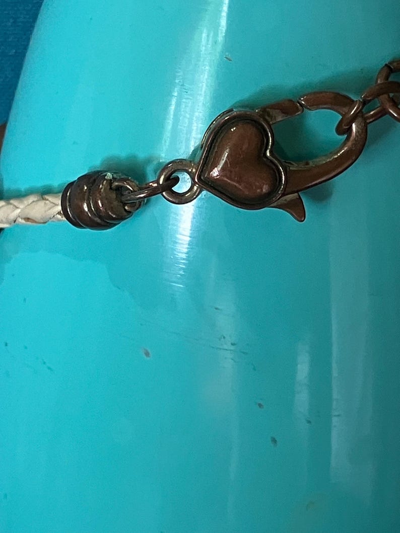 May include: A white braided cord bracelet featuring a metallic bronze-toned lobster claw clasp. The clasp mechanism is accented with a small, smooth, heart-shaped detail in a matching bronze finish, connecting to a cylindrical end cap.