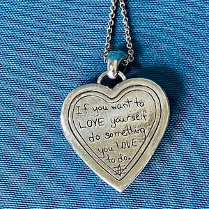 May include: A silver-toned heart-shaped pendant on a matching chain. The pendant features an engraved message that reads: If you want to LOVE yourself do something you LOVE to do. A small symbol is etched at the bottom of the heart-shaped metal piece.