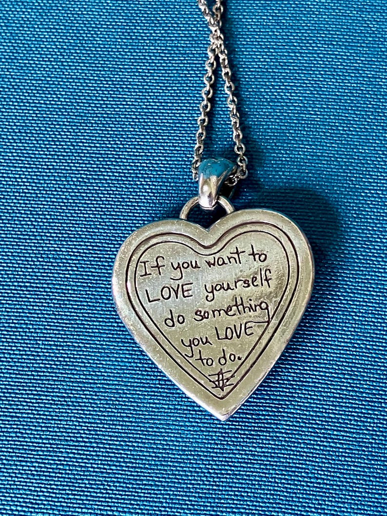 May include: A silver-toned heart-shaped pendant on a matching chain. The pendant features an engraved message that reads: If you want to LOVE yourself do something you LOVE to do. A small symbol is etched at the bottom of the heart-shaped metal piece.