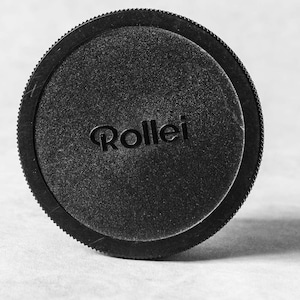 May include: Black camera lens cap with the word "Rollei" printed on it.
