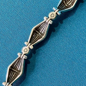 K&ouml;nnte beinhalten: Silberfarbenes Armband mit einem sich wiederholenden Muster aus stilisierten bootsf&ouml;rmigen Gliedern. Jedes Glied ist durch einen kleinen herzf&ouml;rmigen Anh&auml;nger verbunden. Das Armband liegt auf einem strukturierten tealfarbenen Hintergrund.