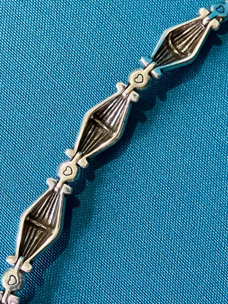 K&ouml;nnte beinhalten: Silberfarbenes Armband mit einem sich wiederholenden Muster aus stilisierten bootsf&ouml;rmigen Gliedern. Jedes Glied ist durch einen kleinen herzf&ouml;rmigen Anh&auml;nger verbunden. Das Armband liegt auf einem strukturierten tealfarbenen Hintergrund.