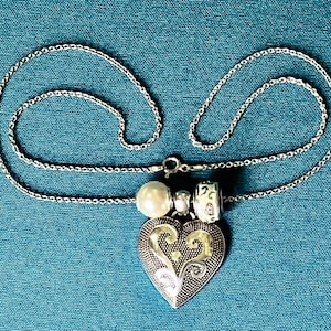 May include: A silver-toned necklace featuring a heart-shaped pendant with an intricate embossed scroll pattern. The pendant is accented by a single white pearl bead and a small silver-toned charm with a decorative etched design on the chain.