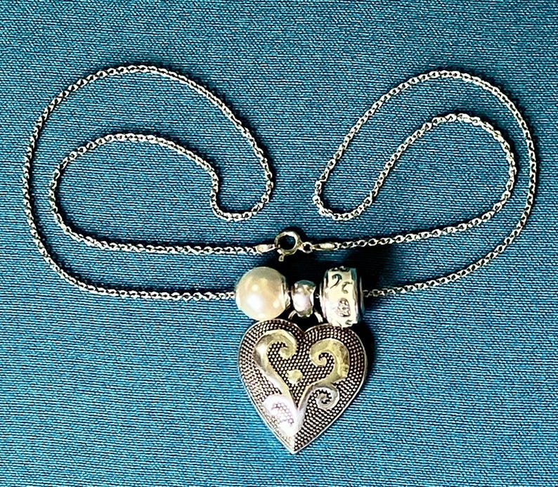 May include: A silver-toned necklace featuring a heart-shaped pendant with an intricate embossed scroll pattern. The pendant is accented by a single white pearl bead and a small silver-toned charm with a decorative etched design on the chain.