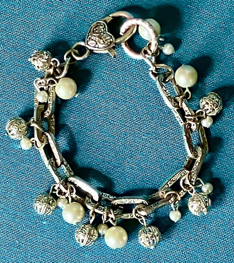 May include: A silver-toned chain link bracelet featuring a heart-shaped clasp and alternating charms. The charms consist of round white faux pearls and small, textured silver-toned spheres. The bracelet links have an engraved pattern.
