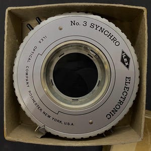 May include: A silver Ilex No. 3 Synchro Electronic camera shutter in its original cardboard box. The shutter has the text "Ilex Optical Company Rochester, New York, U.S.A." and "Electronic" printed on it.