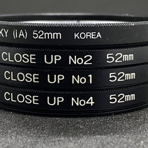 May include: A stack of black camera lens filters. The top filter reads "SKY (IA) 52mm KOREA." Below are three "CLOSE UP" filters, each with "52mm" and "JAPAN" printed on them. The filters are stacked on top of each other.
