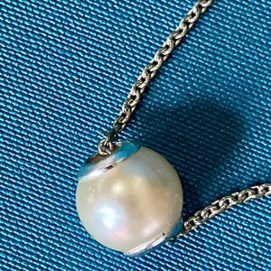 May include: A silver-toned metal chain necklace featuring a single, spherical, off-white pearl pendant. The pearl is held in place by a polished silver-toned metal cap at the top and bottom, creating a clean, simple, and classic jewelry piece.