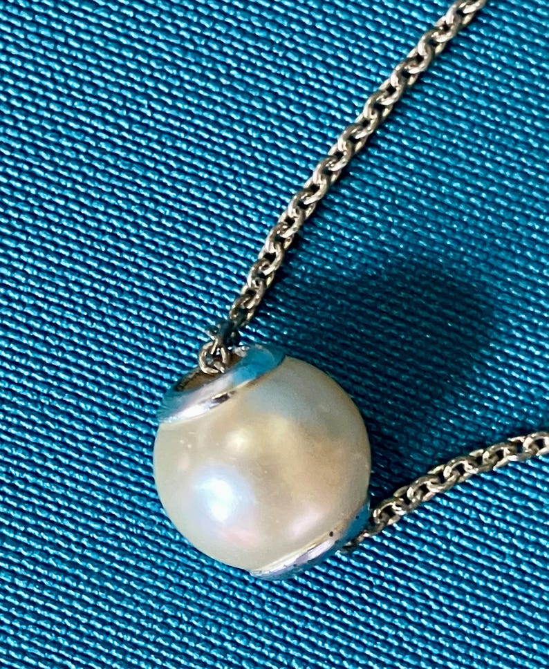 May include: A silver-toned metal chain necklace featuring a single, spherical, off-white pearl pendant. The pearl is held in place by a polished silver-toned metal cap at the top and bottom, creating a clean, simple, and classic jewelry piece.