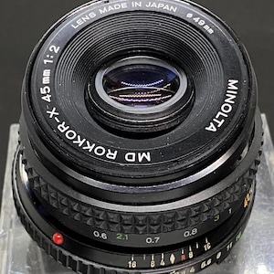 Puede incluir: Un objetivo de cámara negro Minolta MD Rokkor-X 45mm 1:2. El objetivo está marcado con "LENS MADE IN JAPAN" y "49mm". Tiene un anillo de enfoque con marcas de distancia en metros y un anillo de apertura. El objetivo tiene un punto rojo.