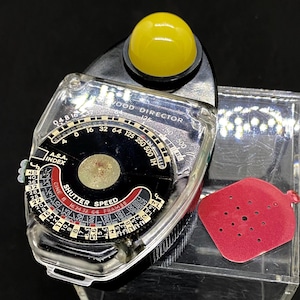 May include: A black and silver light meter with a yellow dome on top. The meter has a dial with the words "WOOD DIRECTOR" and numbers from 4 to 500. The dial also has the words "A.S.A. INDEX" and "SHUTTER SPEED".