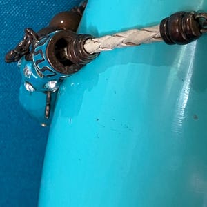 May include: A bracelet featuring a braided cream-colored leather cord, accented with dark metallic beads and a bright turquoise blue charm decorated with intricate black patterns and embedded clear crystals, secured by a metallic clasp mechanism.