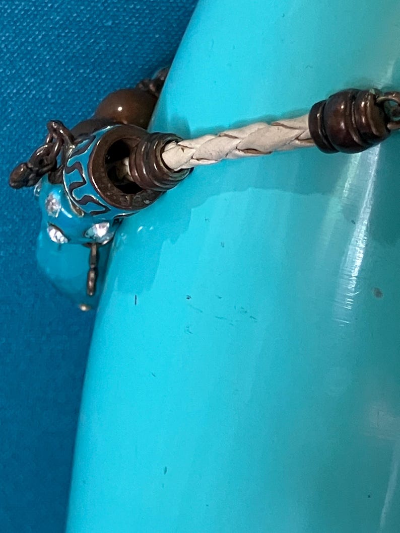 May include: A bracelet featuring a braided cream-colored leather cord, accented with dark metallic beads and a bright turquoise blue charm decorated with intricate black patterns and embedded clear crystals, secured by a metallic clasp mechanism.