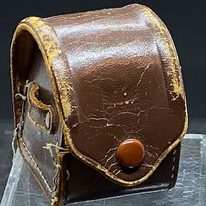 May include: A brown leather case with a flap and a button closure. The case is worn and has a vintage look.