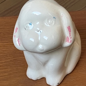 May include: A ceramic figurine of a seated puppy dog. The dog is primarily white with pink accents on the ears and blue eyes. The figurine has a glossy finish and is sitting on a wooden surface.