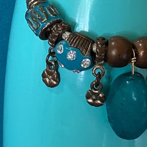May include: A braided white leather cord necklace featuring a variety of beads, including turquoise-colored enamel beads with metallic accents, small bronze-toned flower charms, smooth brown wooden beads, and a large, translucent teal stone pendant.