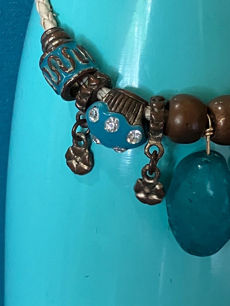 May include: A braided white leather cord necklace featuring a variety of beads, including turquoise-colored enamel beads with metallic accents, small bronze-toned flower charms, smooth brown wooden beads, and a large, translucent teal stone pendant.