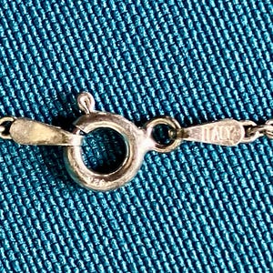 May include: A silver-toned metal chain necklace featuring a circular spring ring clasp. The clasp mechanism is stamped with the text 925 and the word ITALY on the adjacent metal tab, indicating the origin and material composition of the jewelry piece.
