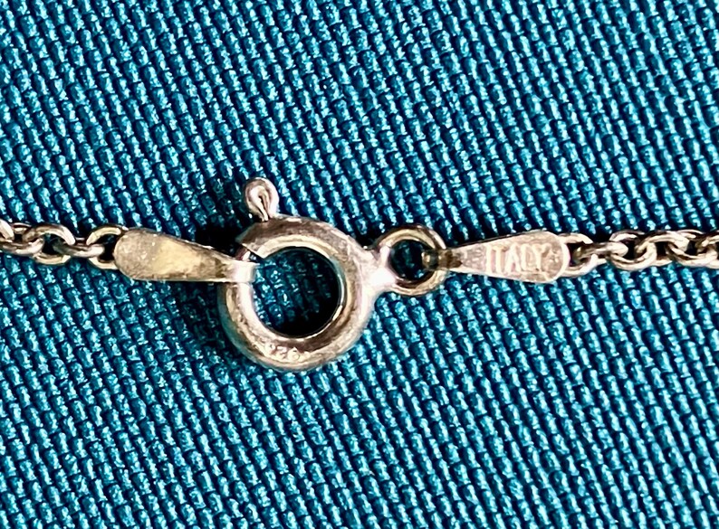 May include: A silver-toned metal chain necklace featuring a circular spring ring clasp. The clasp mechanism is stamped with the text 925 and the word ITALY on the adjacent metal tab, indicating the origin and material composition of the jewelry piece.