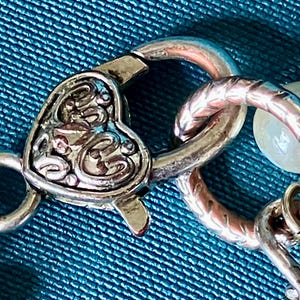 May include: A silver-toned metal lobster claw clasp featuring a heart-shaped design with intricate embossed scrollwork patterns. The clasp is connected to a textured circular jump ring and a portion of a chain with a small white bead visible.