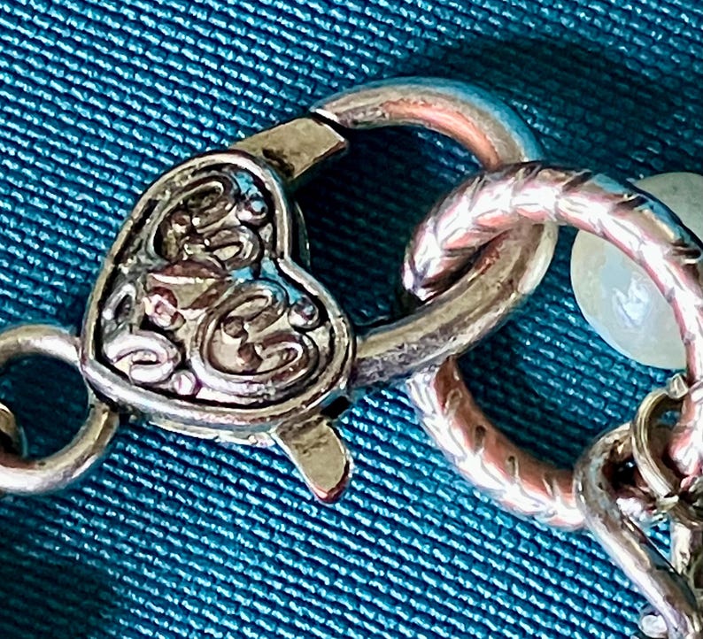 May include: A silver-toned metal lobster claw clasp featuring a heart-shaped design with intricate embossed scrollwork patterns. The clasp is connected to a textured circular jump ring and a portion of a chain with a small white bead visible.